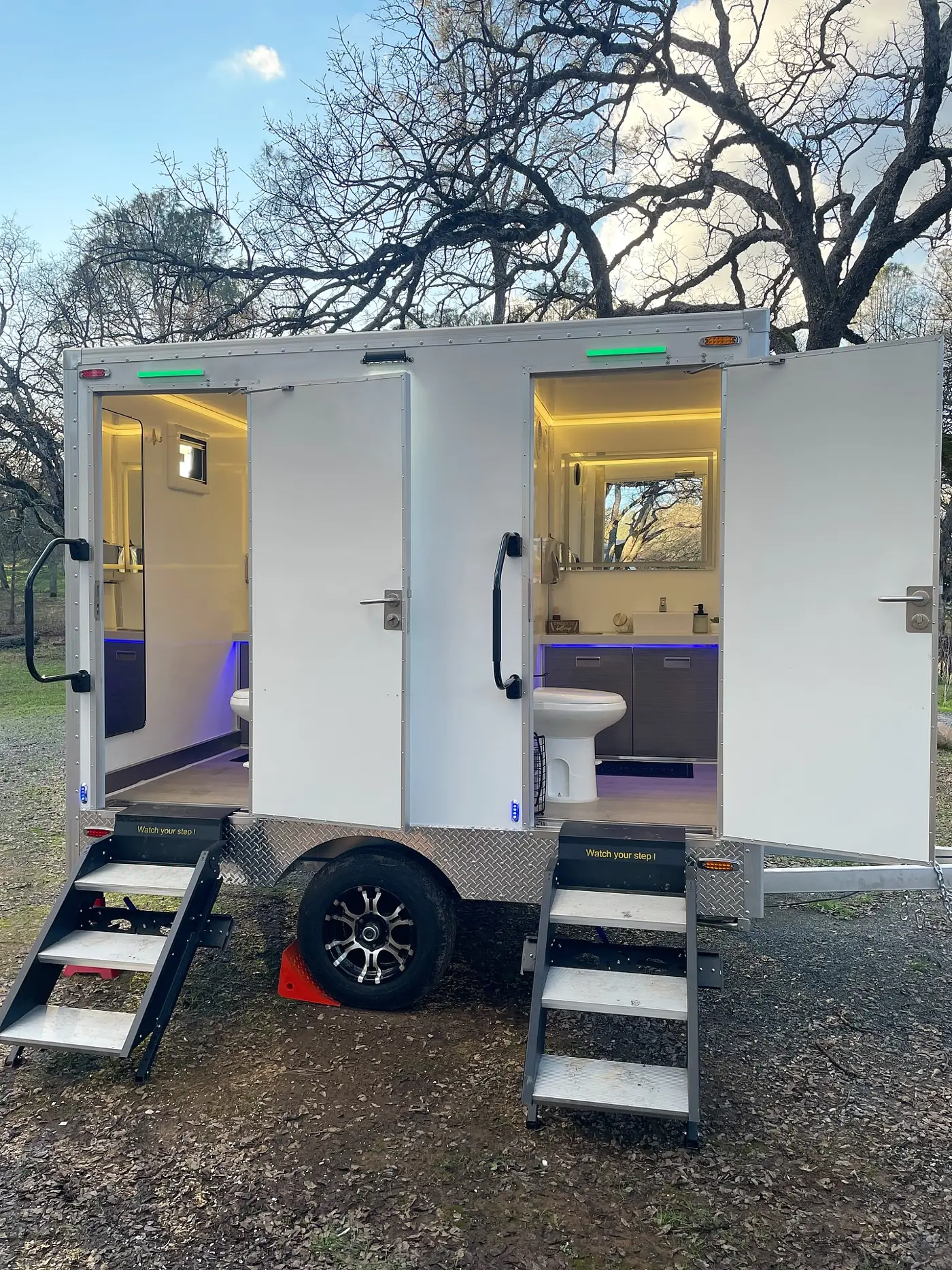 Lit two-station restroom trailer parked on grass at dusk for an outdoor wedding