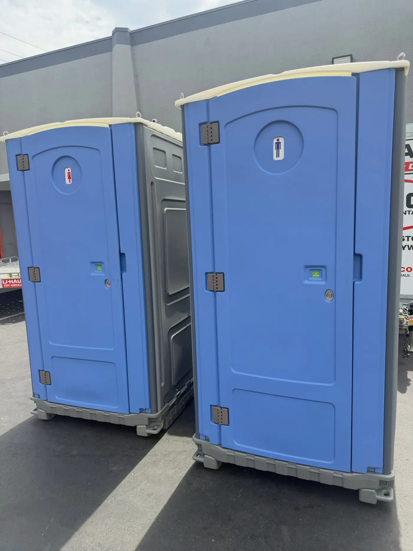 Two blue standard porta potties set side by side on gravel at a job site